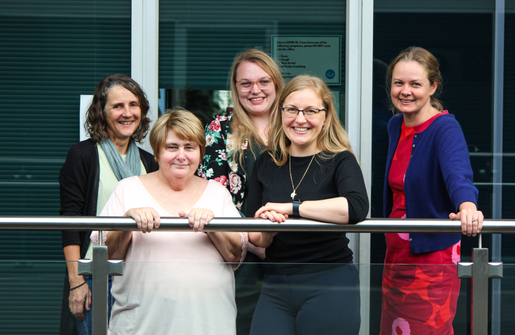 Five women, all white, four with blonde hair and one with brown, are smiling for the camera. This is the core GeneEQUAL team.