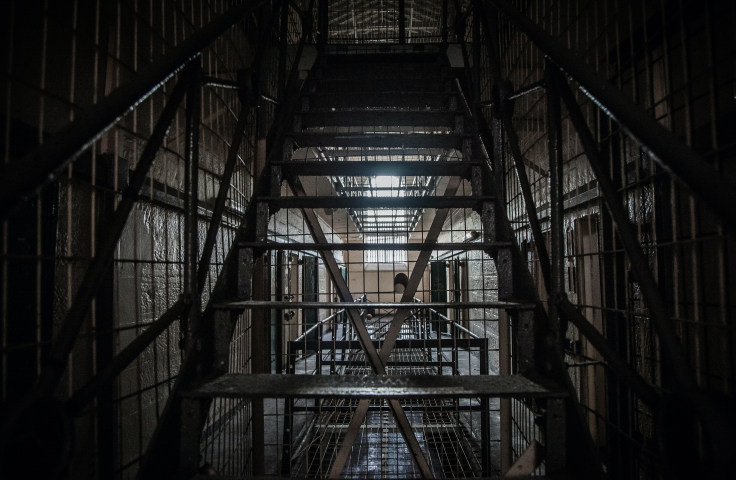 A dark photo of Old Parramatta Gaol, with stairs ascending in the foreground and a stark prison corridor lit by a high window in the background