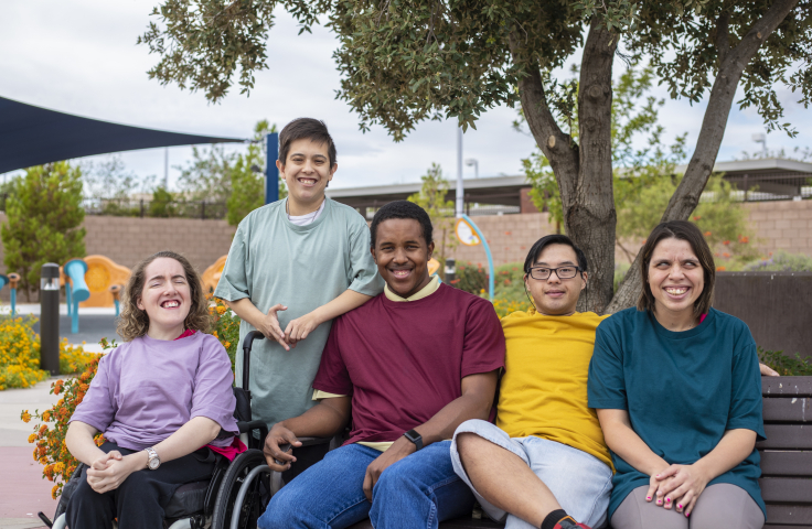 Five young people sitting on a park bench smiling towards the camera 