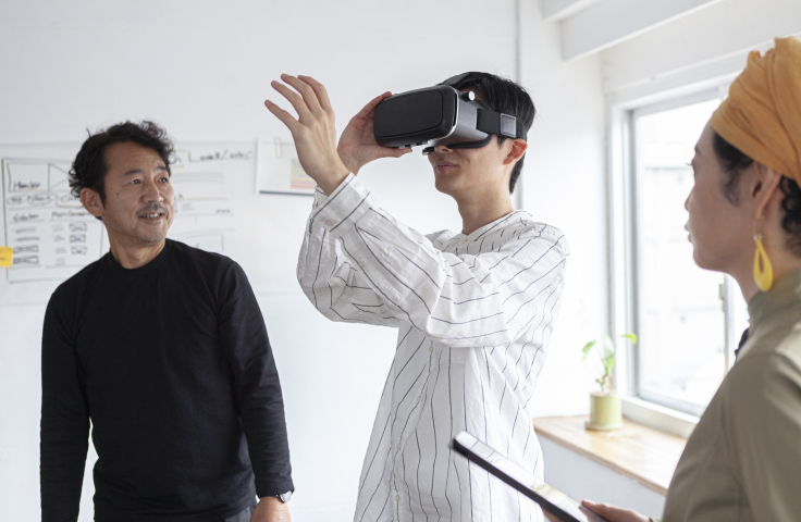 One man looking through a virtual reality headset with two colleagues looking at him. 